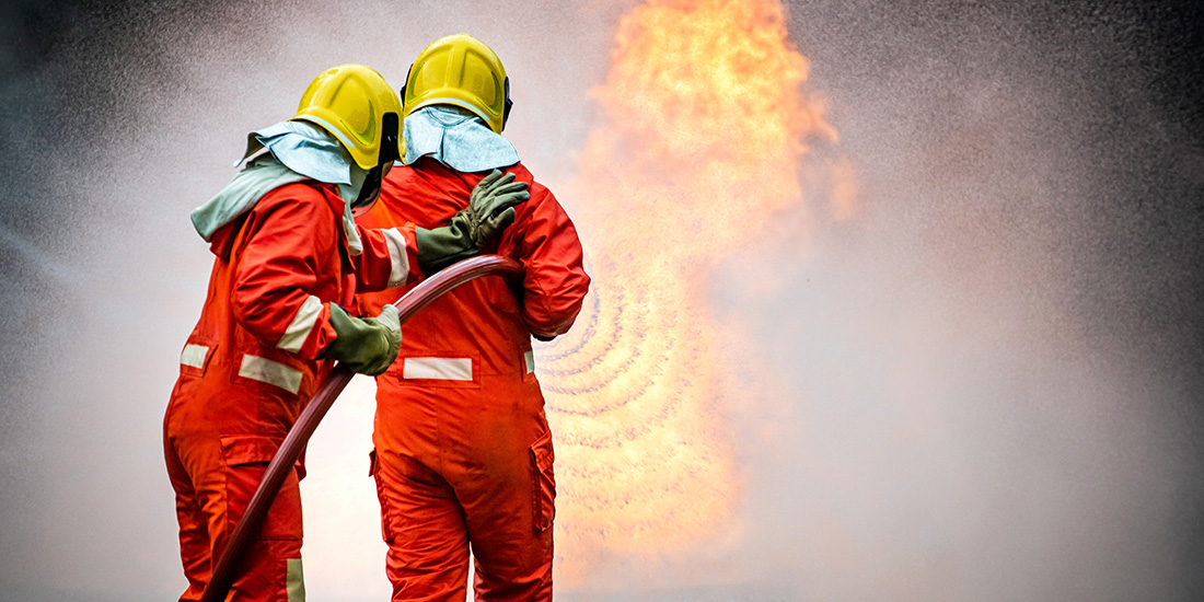 Two firefighters holding a fire hose in front of flames and smoke