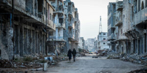 A couple of people walking down a dirty street with rumble from destroyed homes in Daraa, Syria