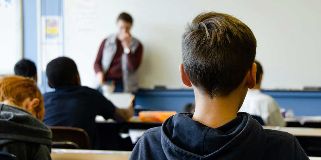 A teacher standing at a white board in the background and the back of a child's head in the foreground
