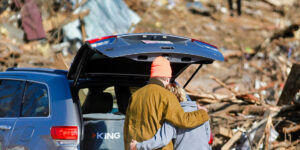 Two people with their arms around each other with a car and rubble from a tornado