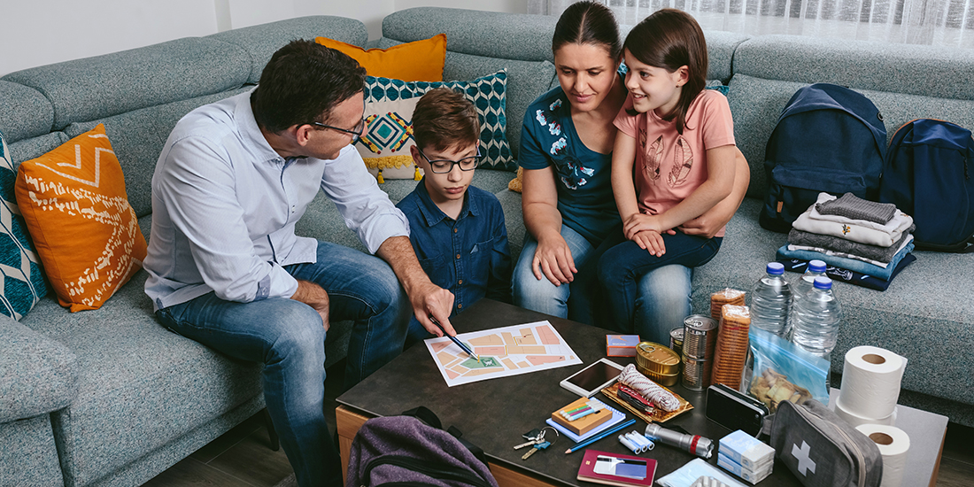 A man and a woman sitting on a sofa with two children looking at a coffee table covered in prepredness supplies: evacuation map, toilet paper, first aid kit, food, water, flashlight, cloths, keys.