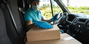 Delivery driver wearing mask and gloves is holding a cellphone looking at cardboard boxes