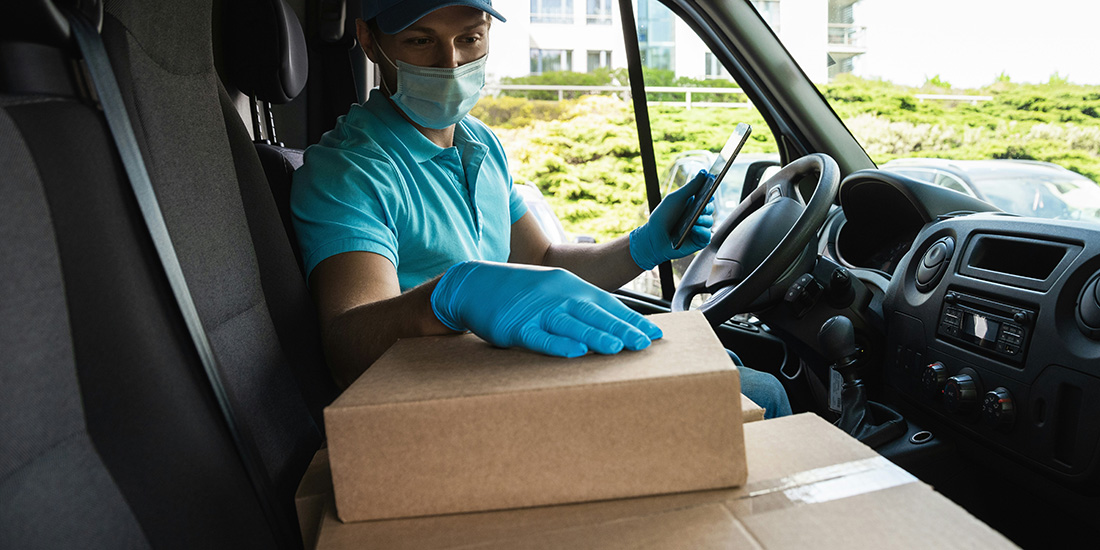 Delivery driver wearing mask and gloves is holding a cellphone looking at cardboard boxes