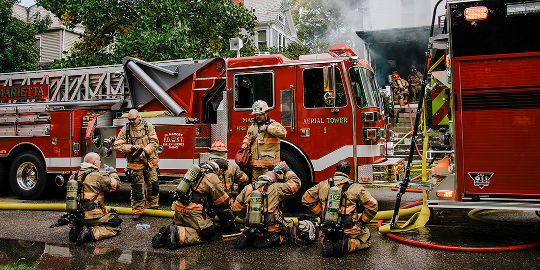 A group of firefighters behind a firetruck drinking water and cooling off, as other firefighters enter a building with smoke