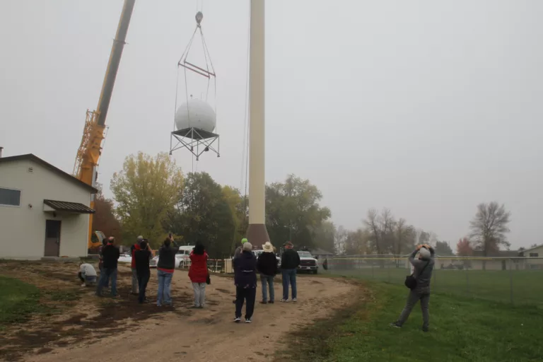 A group of people watching radar equipment being lifted into the air by a large crane.