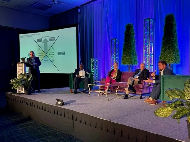 Four panel members sitting on a stage and a moderator standing at a podium.