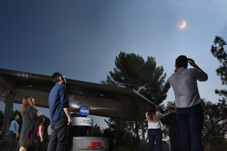 A group of people with eclipse glasses looking up to the sky in front of a NASA welcome sign