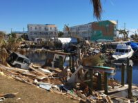 Marina, a fishing boat, and a Jeep are covered in debris. Apartment buildings in the background have damaged roofs and siding.