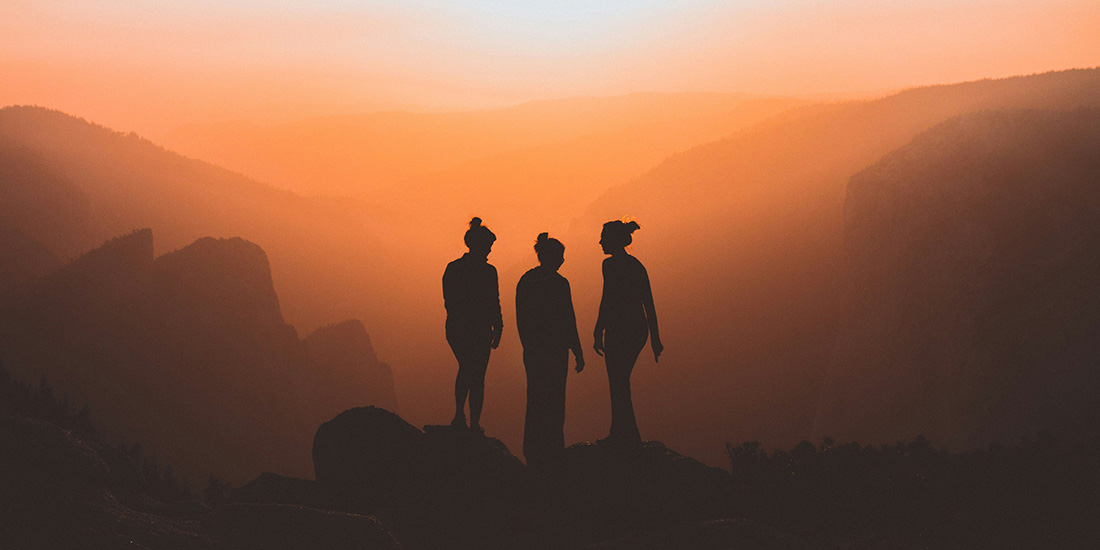 silhouette of three women on mountain cliff with sun setting in background