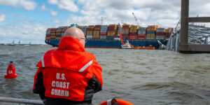 Man in U.S. Coast Guard jacket looks over the water at a cargo ship and bridge that collapsed into the water next to it