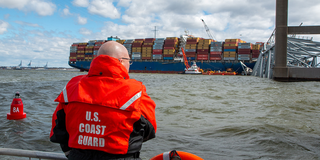 Man in U.S. Coast Guard jacket looks over the water at a cargo ship and bridge that collapsed into the water next to it