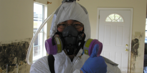 Person wearing glasses, white protective suit, blue gloves, and respirator, holding up thumb in a room with mold on the walls.