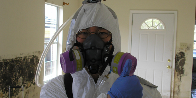 Person wearing glasses, white protective suit, blue gloves, and respirator, holding up thumb in a room with mold on the walls.