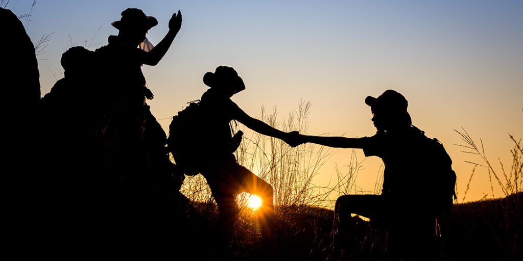 silhouette of Scout students showing help to each other Hold hands and pull each other up from the cliff to explore the beautiful forest in the evening as the sun sets.