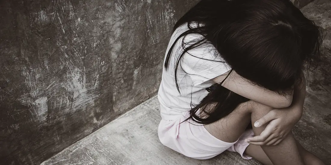 Person sitting curled up on a concrete floor in the corner of a worn room, with arms wrapped around bent knees and head resting on folded arms. The image is shown from above, with textured gray walls and floor creating a stark, somber setting.