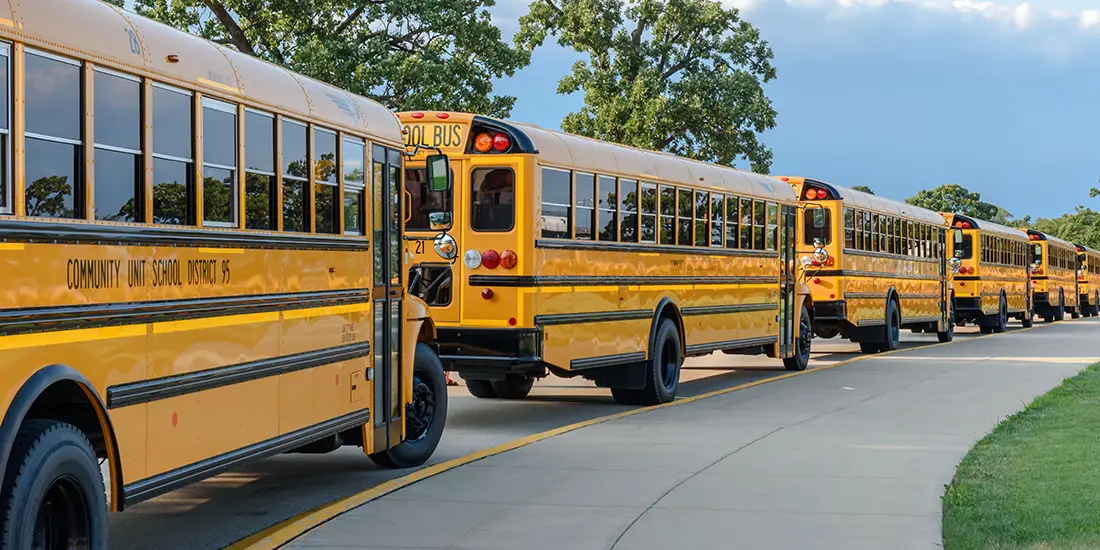 A line of yellow school buses is parked along a curved roadway beside a sidewalk and grassy area. Large trees and a partly cloudy sky are visible in the background. The buses are positioned one behind another, facing the same direction, with the nearest bus shown prominently in the foreground.