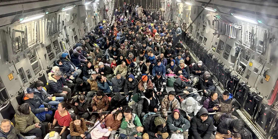 A large military cargo aircraft interior filled with hundreds of evacuees seated closely together on the floor and along the sides of the aircraft. People are bundled in jackets and cold-weather clothing, with bags and personal belongings around them. The aircraft's gray metal walls, equipment panels, and overhead lighting are visible, emphasizing the crowded conditions during a mass transportation or evacuation operation.