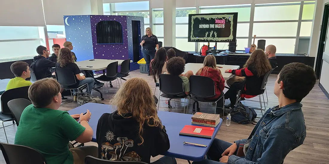 A group of students sits at tables in a classroom or community learning space while a presenter stands at the front of the room beside a display screen. The screen shows the title “Brains Beyond the Brains.” Large windows provide natural light, and educational materials, chairs, and tables are arranged throughout the room for an interactive presentation or workshop.