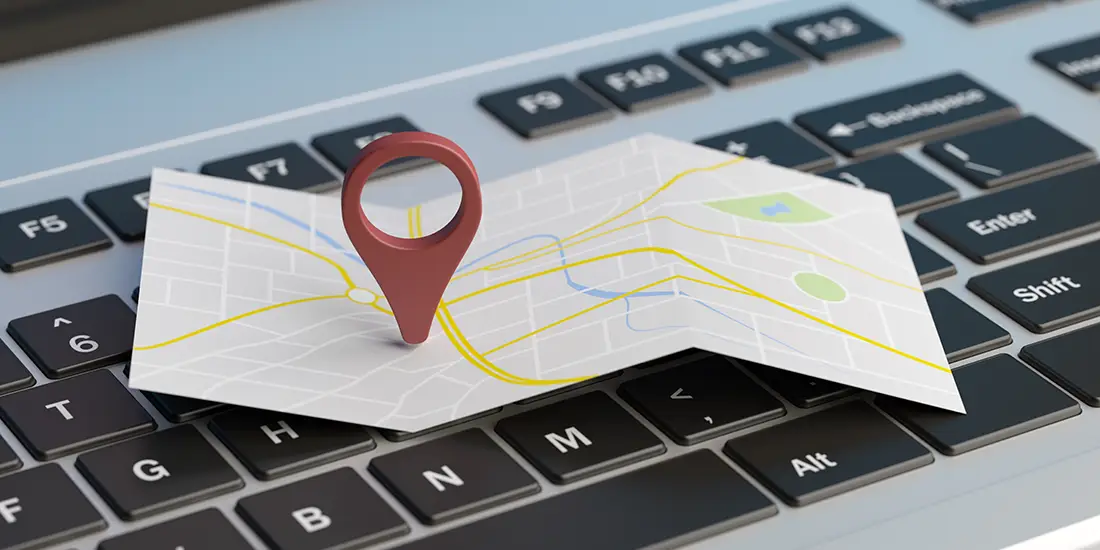 Folded street map with a red location pin placed on top of a laptop keyboard. The map displays roads, intersections, and geographic features in a simplified digital style, while the keyboard keys are visible beneath it.