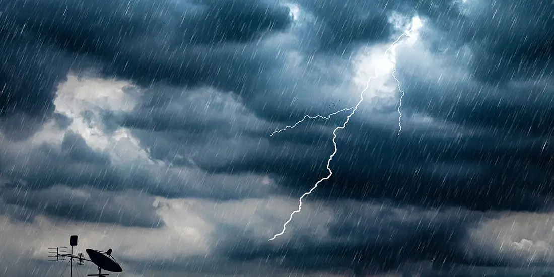 Dark storm clouds fill the sky as heavy rain falls across the scene. A bright lightning bolt streaks through the clouds above silhouetted rooftop antennas and a satellite dish, illustrating severe weather and hazardous storm conditions.