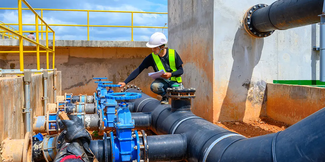 Worker wearing a hard hat and high-visibility safety vest inspecting large water utility pipes and valve assemblies at an industrial facility. The person is kneeling beside interconnected black and blue pipelines while holding documents, with concrete structures, safety railings, and additional piping visible throughout the site. The image depicts maintenance and inspection activities associated with water infrastructure systems.