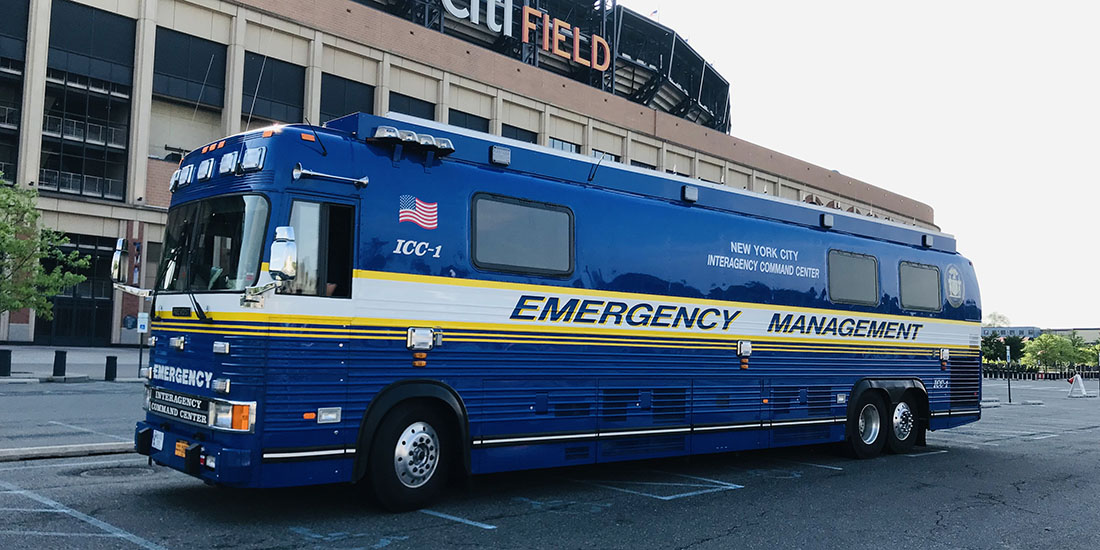 Large blue mobile command vehicle labeled “Emergency Management” and “New York City Interagency Command Center” parked outside Citi Field. The specialized emergency management vehicle features multiple windows, rooftop communications equipment, and yellow striping along the sides. The stadium structure is visible in the background, with part of the sign displayed above the vehicle. The image depicts a mobile command and coordination asset used for emergency management operations and incident response.