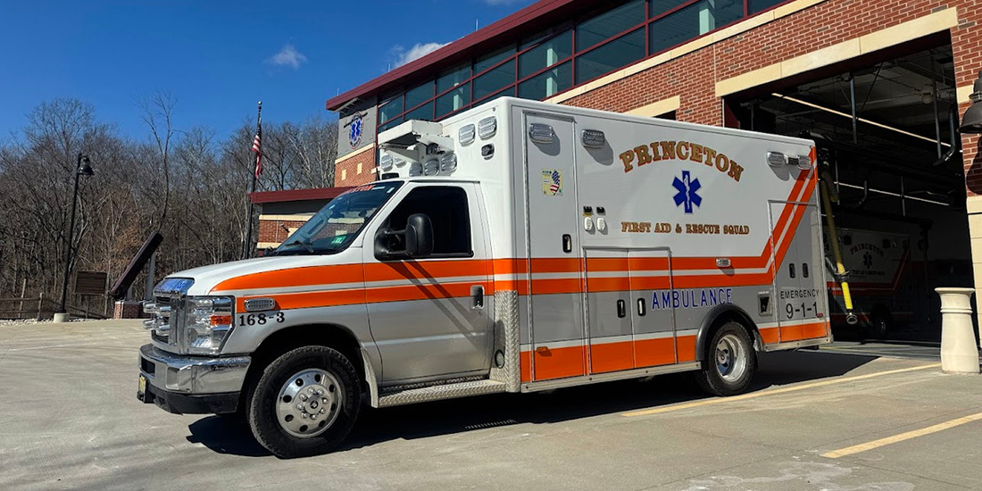 Orange-and-white ambulance marked “Princeton First Aid & Rescue Squad” parked outside a brick emergency services building on a clear day. The ambulance is positioned in front of an open vehicle bay, with a second emergency vehicle visible inside the station. An American flag stands near the building, and leafless trees line the background under a blue sky with scattered clouds.
